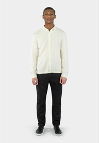 Crème knoopshirt van een textuurstof, gecombineerd met zwarte broek en zwarte sneakers. Eenvoudig ontwerp, klassieke pasvorm en neutrale kleuren.