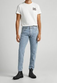 Vit T-shirt med korta ärmar och "Pepe Jeans"-logotyp, kombinerad med blå och vita randiga jeans och svarta ankelboots. Enkel design, ledig passform.