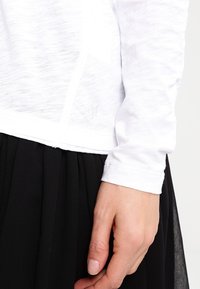 En närbild av en kvinnas hand som håller en svart kjol, iförd en vit långärmad topp. Fokuset ligger på kläderna och handens position.