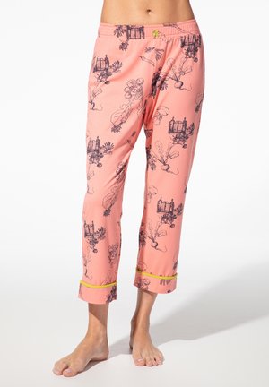 Rosa gekürzte Hose mit schwarzen botanischen und Käfigmustern, gelber Besatz an den Bündchen, getragen von einer barfuß stehenden Person vor weißem Hintergrund.