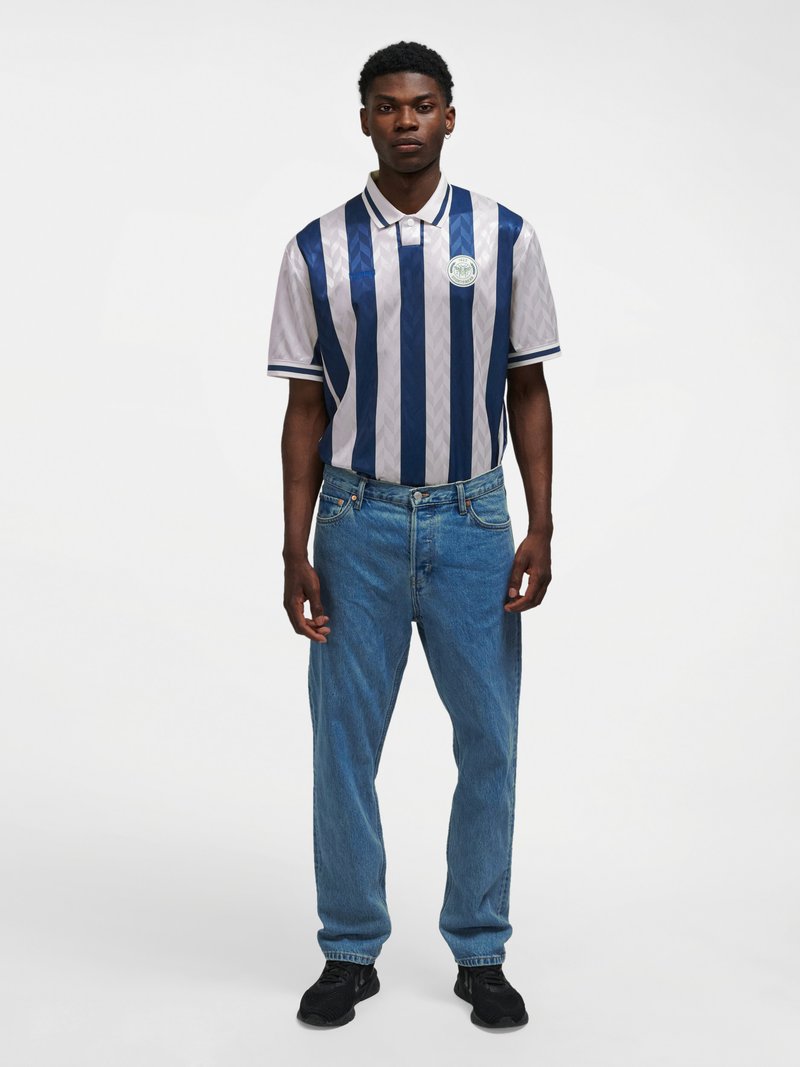 Un homme debout portant un polo à rayures verticales bleues et blanches, un jean bleu et des baskets noires sur fond blanc uni.