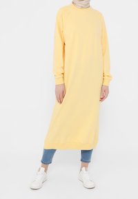 Vestido sudadera amarillo de gran tamaño con mangas raglán largas, puños y dobladillo acanalados. Combinado con jeans cortos en azul claro y zapatillas blancas.