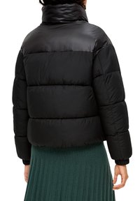 Schwarze Pufferjacke mit hohem Kragen, mit einem gesteppten Design, glänzender Textur und elastischen Bündchen. Über einem grünen Ripprock getragen.