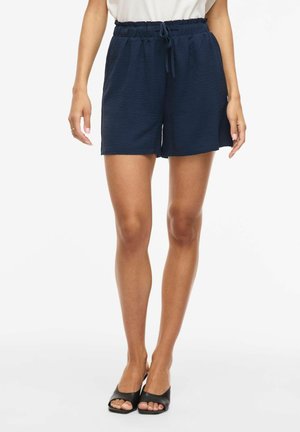 Naine, kes kannab tekstuuriga tumelikke siniseid lühikesi pükse, millel on elastne vöökoht ja pael, koos mustade avatud varbaga kontsakingadega, seisab valge tausta ees.