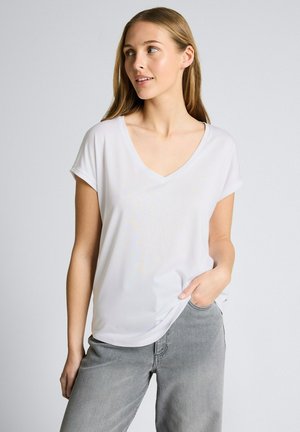 Junge Frau mit langen hellen Haaren, die ein weißes V-Ausschnitt-T-Shirt und graue Jeans trägt, steht mit einer Hand in der Tasche und schaut nach rechts.