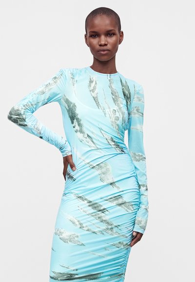 Femme aux cheveux courts portant une robe bleu clair à manches longues froncée avec des motifs abstraits de coups de pinceau gris, posant avec une main sur la hanche.