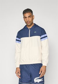 Marineblauwe en crèmekleurige rits hoodie met elastische tailleband, met horizontale blauwe strepen op de mouwen en een klein groen logo.