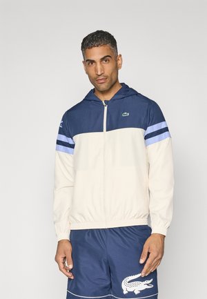 Giacca a zip con cappuccio in blu navy e crema, con vita elastica, caratterizzata da strisce orizzontali blu sulle maniche e un piccolo logo verde.