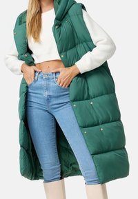 Gilet imbottito verde con cappuccio, dotato di bottoni a pressione dorati e tessuto trapuntato, indossato sopra un maglione bianco e jeans blu chiaro.