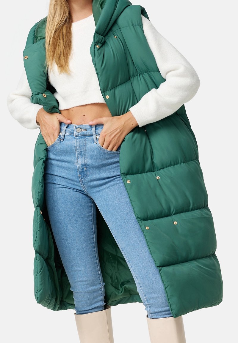 Gilet imbottito verde con cappuccio, dotato di bottoni a pressione dorati e tessuto trapuntato, indossato sopra un maglione bianco e jeans blu chiaro.
