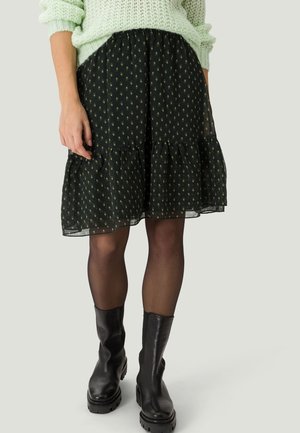 Femme portant une jupe noire à motifs jusqu’aux genoux, des collants noirs transparents, des bottes noires épaisses et un pull en maille vert clair avec les manches retroussées.