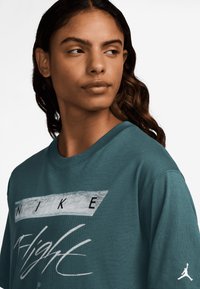 Svetlo turkizna bombažna majica z grafičnim dizajnom v sivi in črni barvi, ki vsebuje besedilo 'NIKE' in 'Flight'. Na rokavu je tudi bel Jumpman logotip.