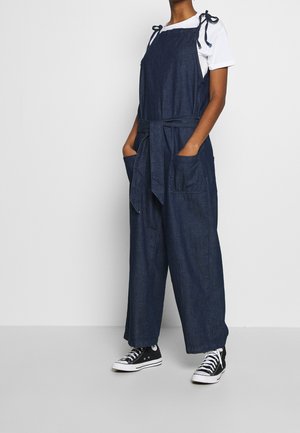Dżinsowy kombinezon w ciemnoniebieskim kolorze z wiązanym paskiem w talii, regulowanymi ramiączkami i dwiema przednimi kieszeniami. Połączony z białą koszulką i czarnymi trampkami.