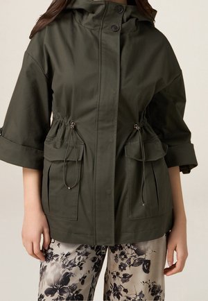 Zielona oliwkowa kurtka z kapturem, przednimi kieszeniami, sznurkami ściągającymi w talii i podwiniętymi rękawami, noszona na spodniach w kwiatowy wzór.