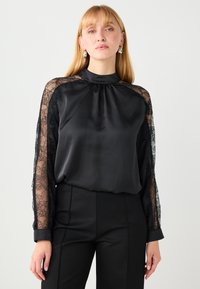Schwarze Satinbluse mit langen Spitzenärmeln, hohem Kragen und gerafftem Ausschnitt. Kombiniert mit schwarzen, maßgeschneiderten Hosen. Elegante Textur und Design-Details.