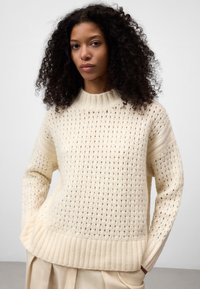Crème gebreide trui met een losse weefpatroon, geribbelde manchetten en een hoge kraag. Zachte textuur en een relaxed fit.