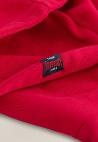 Červená bavlněná mikina s měkkou texturou, se černou nálepkou s nápisem "Superdry" v červeném písmu a bílými detaily, částečně složená.