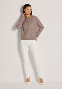 Vrouw in een bruine kabelgebreide trui, witte strakke broek en witte puntige hakken, staande tegen een effen lichte achtergrond.