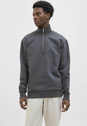 Uomo con felpa grigia con mezza zip e pantaloni beige, in piedi e che guarda di lato contro uno sfondo chiaro e uniforme.