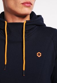 Marinblå sweatshirt med hög krage, försedda med orange dragband och en logo i hexagonal form på bröstet. Mjuk, texturerad tyg.