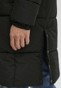 Svart pufferjakke med quiltet tekstur. Har elastiske mansjetter og trykknapper langs sidesømmen. Synlig hånd som griper i stoffet.