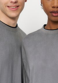 Δύο άτομα που φορούν ίδια γκρι μπλουζάκια με στρογγυλή λαιμόκοψη, εμφανίζοντας το μισό πρόσωπο και το πάνω μέρος του κορμού τους πάνω σε λευκό φόντο.