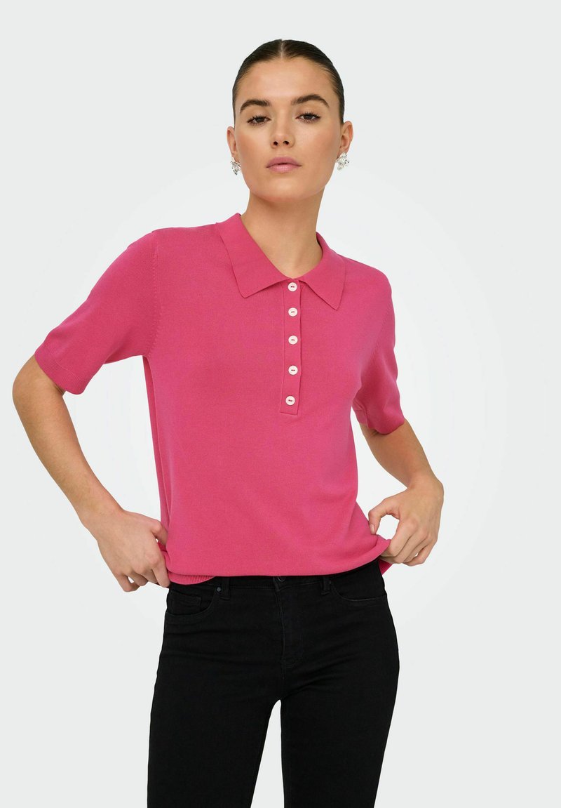 Donna che indossa una maglietta polo corta rosa brillante con bottoni bianchi, pantaloni neri e orecchini floreali su uno sfondo chiaro e semplice.