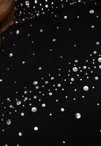 Czarny materiał z rozrzuconymi, przezroczystymi cyrkoniami o różnej wielkości, tworzącymi błyszczący efekt. Teksturowany materiał o gładkim wykończeniu.