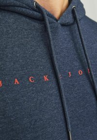 Sudadera azul marino de tejido suave y texturizado, con un bolsillo canguro en la parte delantera, cordones y letras rojas impresas que dicen "JACK" en el pecho.
