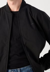 Schwarze Bomberjacke mit Reißverschluss und geripptem Kragen. Glatter Stoff und dezentes Calvin Klein-Logo auf der Brust. Kombiniert mit einem schwarzen Top.