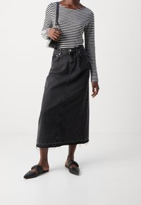 Schwarzer Denim-Maxi-Rock mit fransigem Saum, kombiniert mit einem schwarz-weiß gestreiften Langarmoberteil und schwarzen flachen Schuhen. Kompakte Handtasche.
