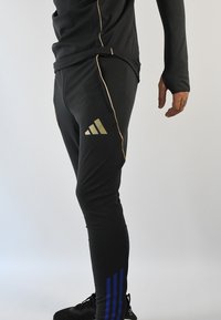 Pantalones deportivos negros con detalles dorados, que presentan un corte ajustado, bolsillos laterales y un diseño de tres rayas azules cerca del tobillo. Material sedoso.