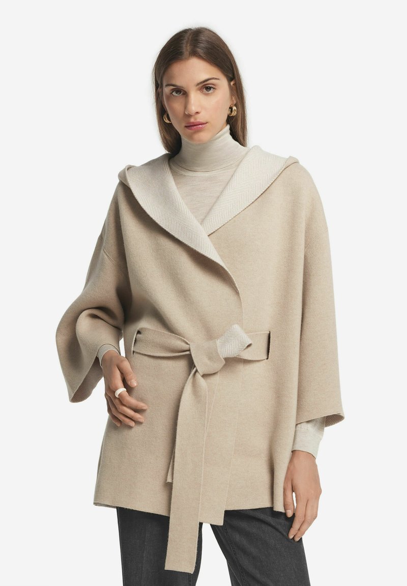 Beige ullblandad ytterklädnad med en bred huva med fiskbensmönster, bälte i midjan och vida ärmar, lagrad över en ljus polotröja.