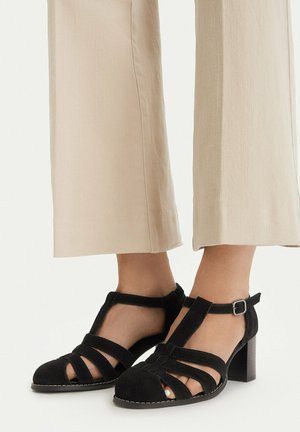 Stopy w czarnych zamszowych sandałach na blokowym obcasie z paskiem typu T i klamrą przy kostce, w połączeniu z beżowymi spodenkami o kroju capri na białym tle.
