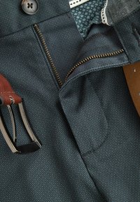 Pantalons pour hommes en tissu sombre avec un motif texturé, dotés d'une fermeture éclair métallique, d'une ceinture en cuir marron et d'une doublure intérieure détaillée.
