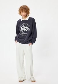 Sudadera marina con texto blanco y gráfico de caballo, combinada con pantalones anchos blancos. Tela de mezcla de algodón, corte relajado y cuello en contraste.