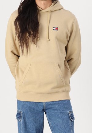 Osoba ubrana w beżową bluzę z kapturem Tommy Jeans z przednią kieszenią i niebieskie spodnie cargo, stojąca z rękami za plecami.