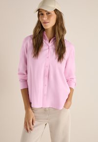 Femme portant une chemise rose pâle boutonnée, un pantalon beige et une casquette de baseball crème, debout avec une main dans la poche devant un fond uni.