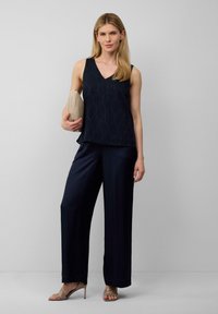 Haut navy à col en V avec un motif ondulé texturé, associé à un pantalon large navy. Le modèle porte une pochette beige et des talons beiges à brides.