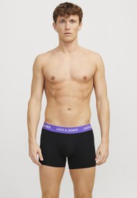 Schwarze Boxer-Shorts mit einem lila elastischen Bund, der das Logo "JACK & JONES" trägt. Der Stoff scheint glatt und eng anliegend zu sein. Das Modell ist oberkörperfrei.