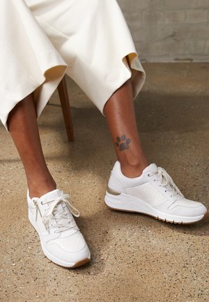 Osoba nosząca białe trampki i kremowe spodnie, siedząca i pokazująca tatuaż na kostce w kształcie czarnego odcisku łapy na prawej nodze.