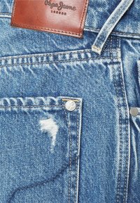 Jeans de denim azul claro com detalhes desgastados, um pequeno bolso frontal, ferragens prateadas e um patch de couro castanho na cintura com a marca.