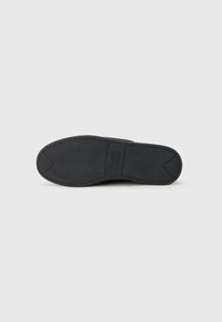 Semelle de chaussure en caoutchouc noir avec une surface texturée, un design légèrement incurvé, un devant arrondi et un logo de marque visible au centre.