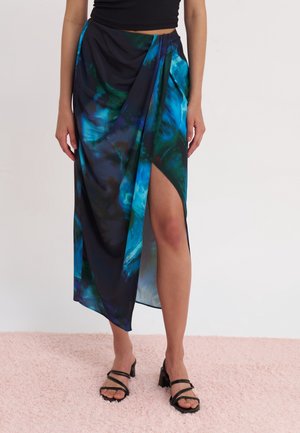 Femme portant une jupe portefeuille abstraite bleue et noire avec une fente haute sur la cuisse, associée à des sandales à talons noires à brides et un crop top noir.