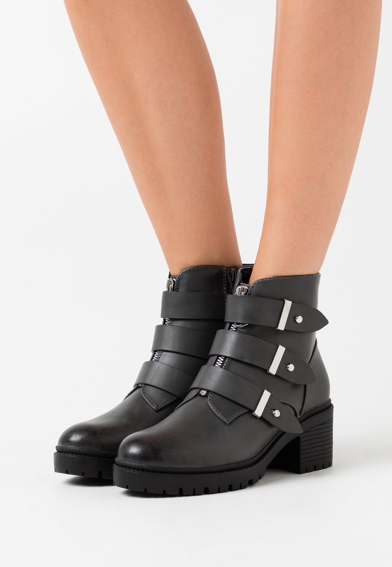 Bottines en cuir noir avec un talon chunky, trois larges sangles avec des éléments en argent, et une fermeture éclair sur le côté. Surface texturée et bout rond.