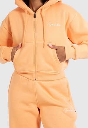 Mujer con sudadera con capucha con cremallera color melocotón y pantalones deportivos a juego, manos en los bolsillos de la sudadera, logo en escritura blanca en el pecho y el muslo.
