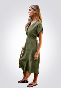 Vestido verde oliva, hasta las rodillas, en tejido texturizado con mangas cortas, escote en V y bolsillos laterales. Lleva sandalias negras de dedo y sostiene un bolso trenzado.
