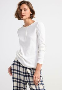 Vit långärmad skjorta med rund halsringning och tre knappar, tillsammans med rutiga, blå och gräddfärgade pyjamasbyxor. Mjuk, ribbad textur.