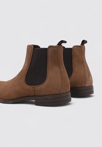 Botas Chelsea de ante marrón con paneles laterales elásticos, un lazo en la parte posterior y una suela de goma plana con detalles texturizados.