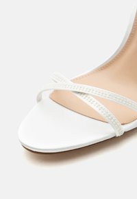 Λευκό ανοιχτό πέδιλο με λεπτούς σταυρωτούς ιμάντες με μικρές υφασμάτινες χάντρες και μπεζ εσωτερική σόλα.